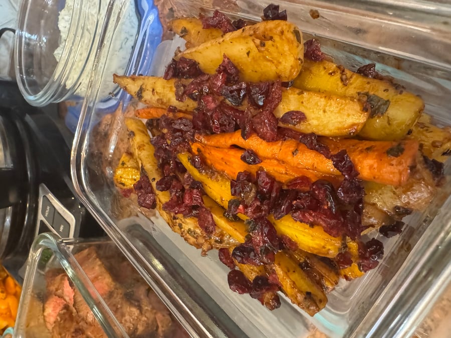 A glass container filled with balsamic cranberry baked carrots, featuring golden roasted root vegetables topped with dark, sweet dried cranberries and savory herbs.
