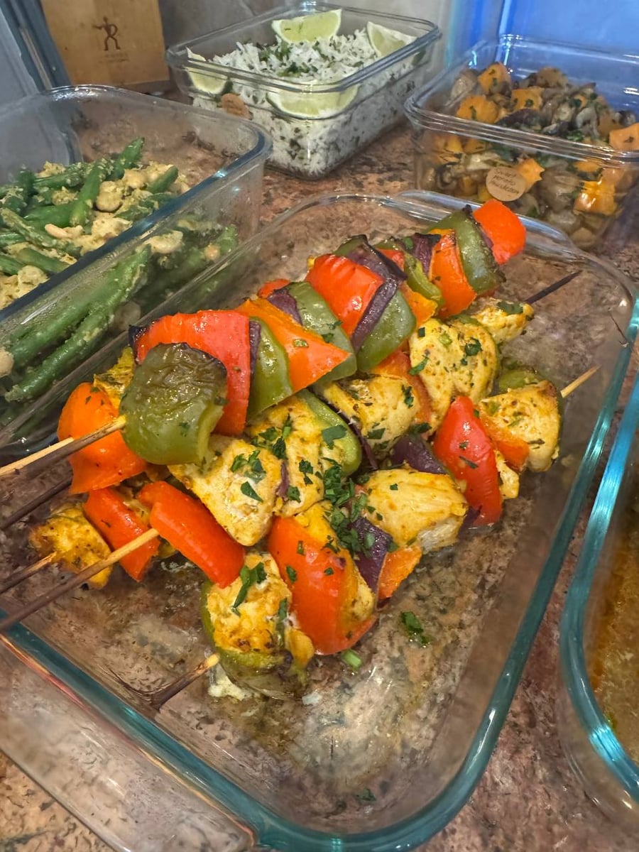 Glass container holding grilled chicken breast and bell pepper skewers with pesto-style green beans, roasted sweet potatoes, and lime cilantro rice nearby.
