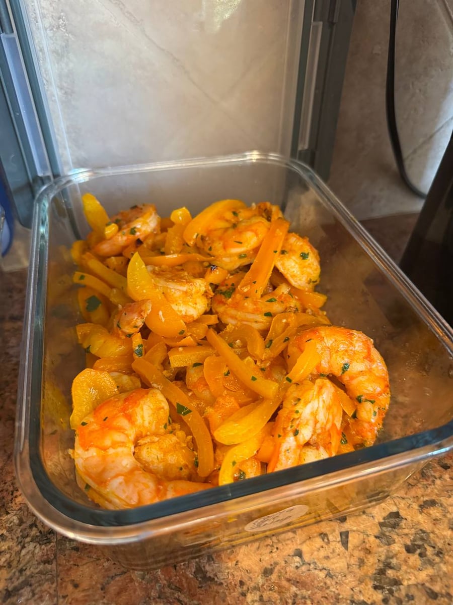 A glass container packed with fajita-style baked shrimp and vibrant yellow bell pepper strips, seasoned and garnished with fresh green herbs.