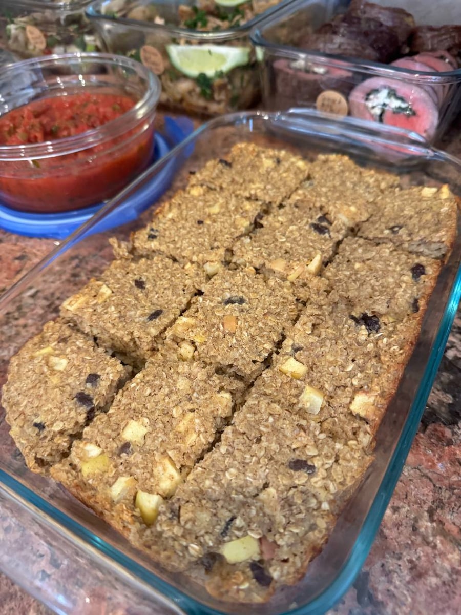 A glass tray filled with rectangular slices of apple cinnamon raisin baked oats, with containers of salsa, chicken, and stuffed steak behind.