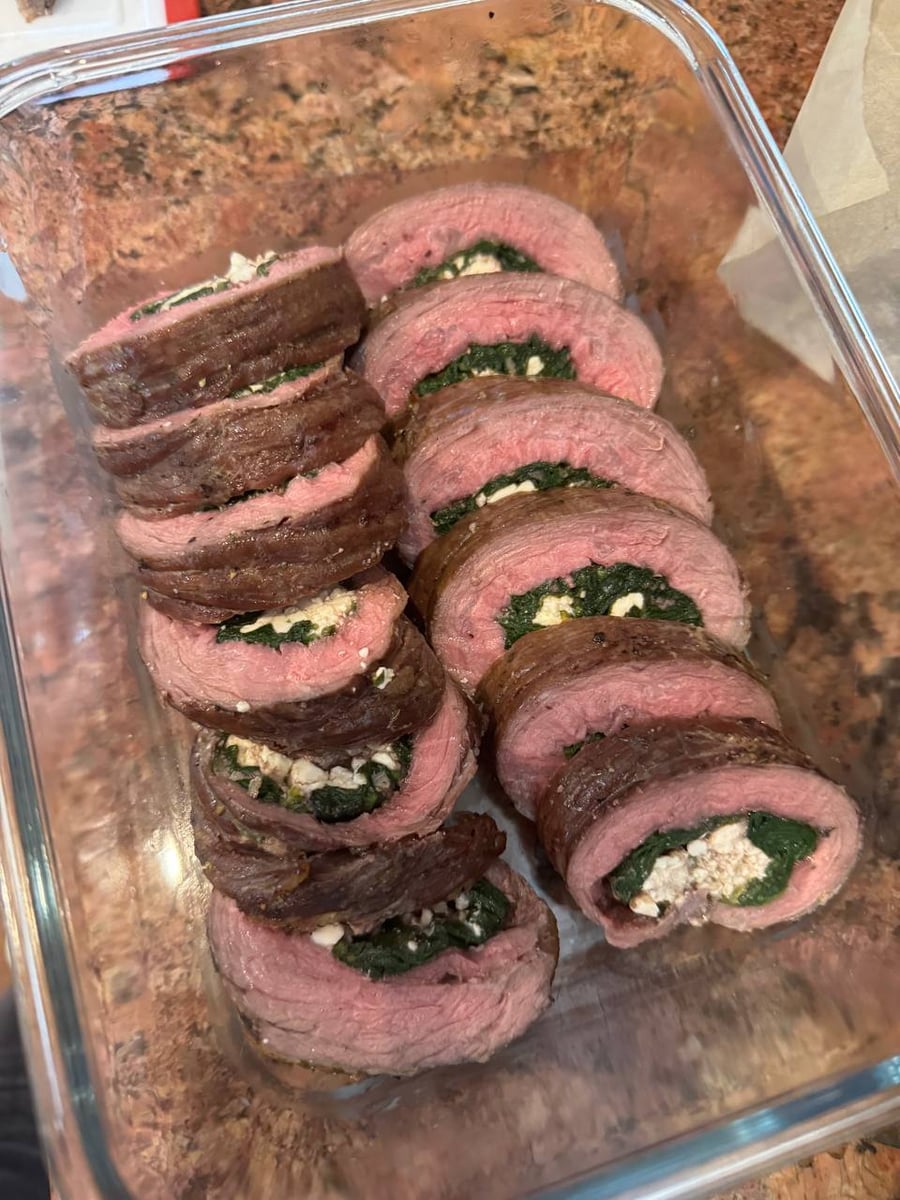 A glass storage container holding neatly arranged, sliced pinwheels of flank steak stuffed with vibrant green spinach and creamy white feta cheese.