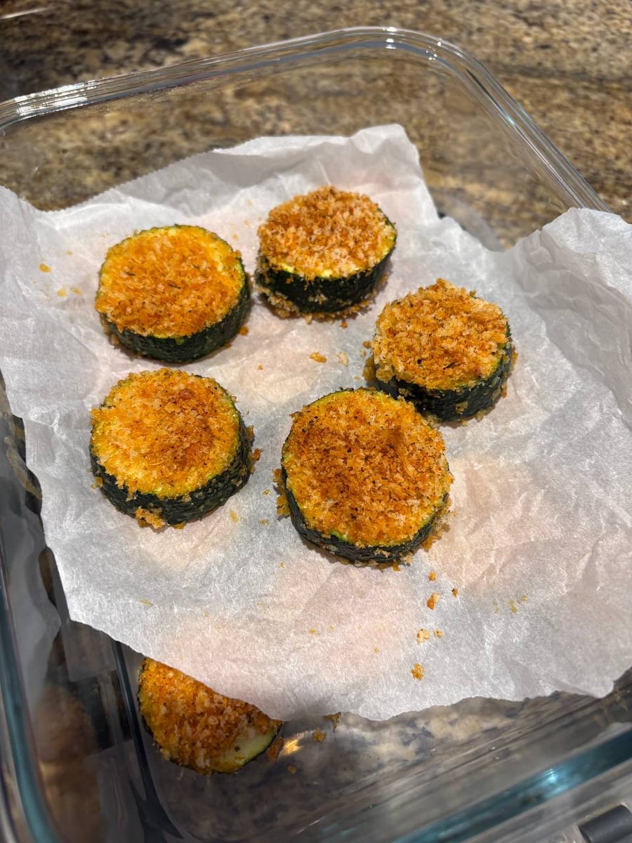 A glass container lined with parchment paper holds five crispy baked zucchini rounds featuring a golden-brown breaded crust and green zucchini centers.