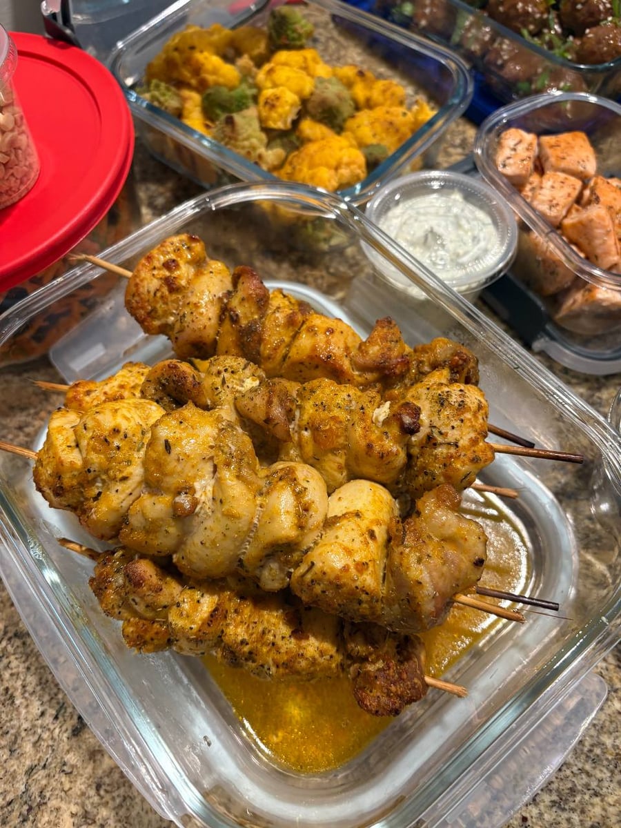 A glass container holds several garlic parmesan chicken skewers resting in a savory sauce alongside a small side of herbed yogurt.