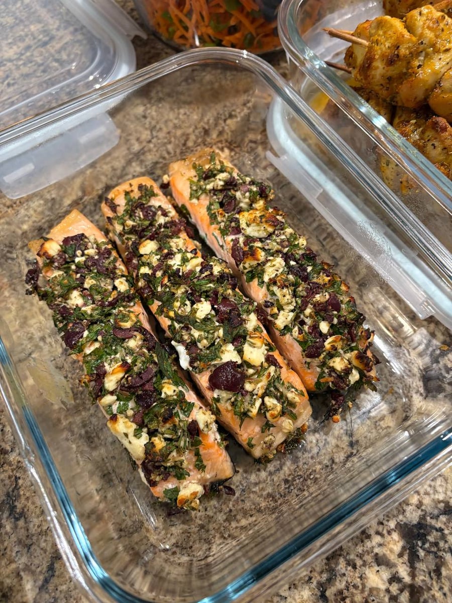 Three roasted salmon fillets topped with crumbled feta cheese, fresh herbs, and chopped kalamata olives, neatly arranged in a clear glass container.