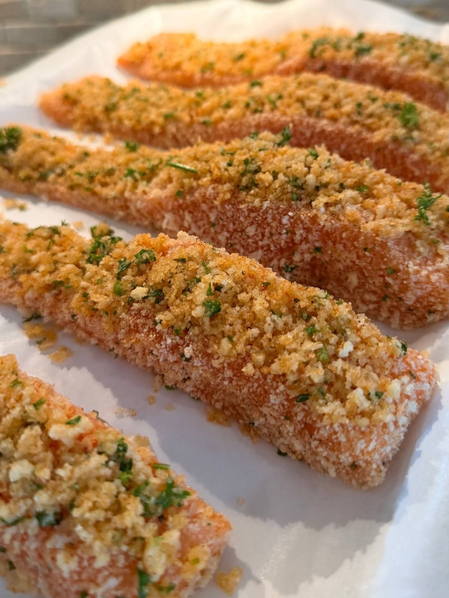 Herb speckled panko sits raw on salmon fillets, parchment underneath and pale brick behind them.