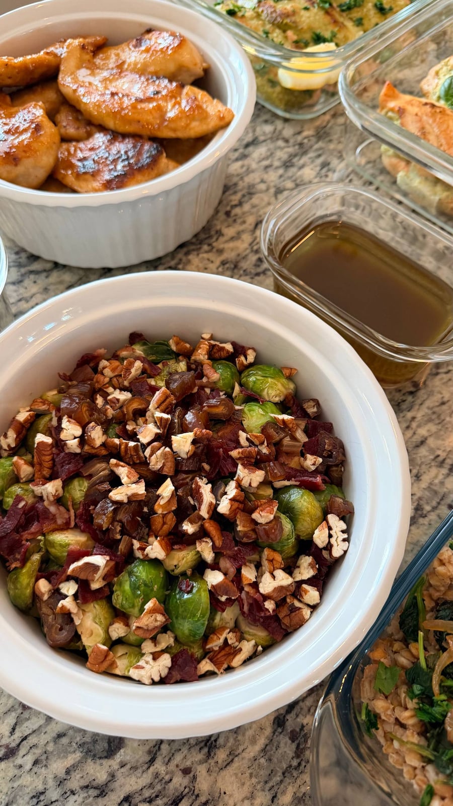 Dates and toasted pecans blanket small brussels sprouts in a white bowl, glazed chicken sitting above.