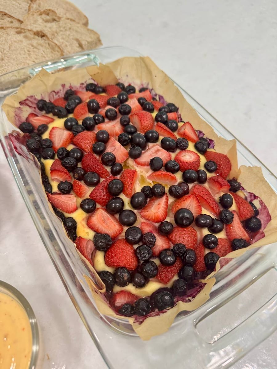 Strawberry halves and blueberries stud the baked yogurt bun, its parchment stained purple at the edge.