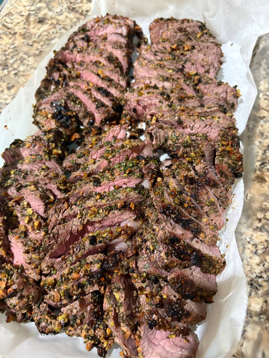 A tray of sliced mint-pistachio crusted grilled lamb chops, featuring succulent medium-rare meat and a golden herb-nut crust served on parchment paper.