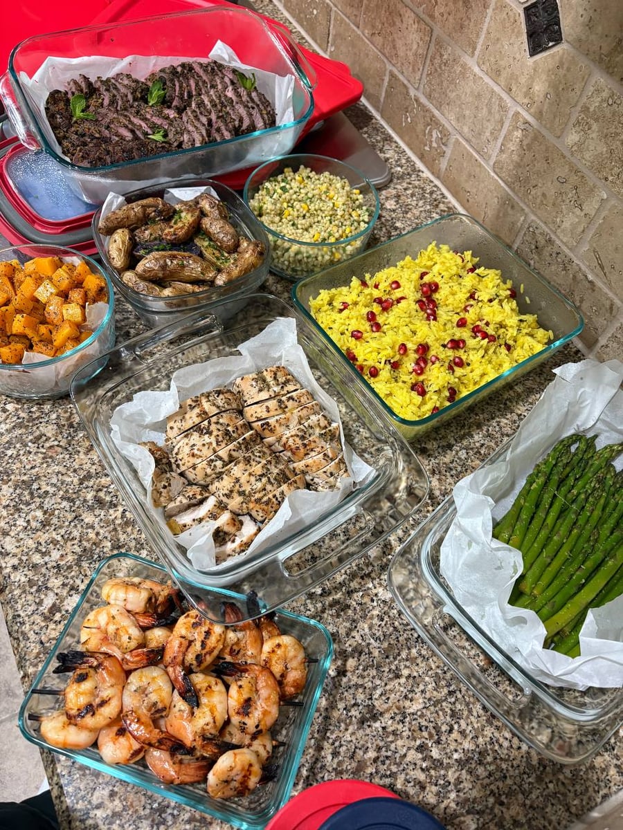 A variety of glass containers holding sliced mint-pistachio lamb, ginger-pear turkey, grilled shrimp, saffron rice, pearl couscous, roasted potatoes, and blanched asparagus.