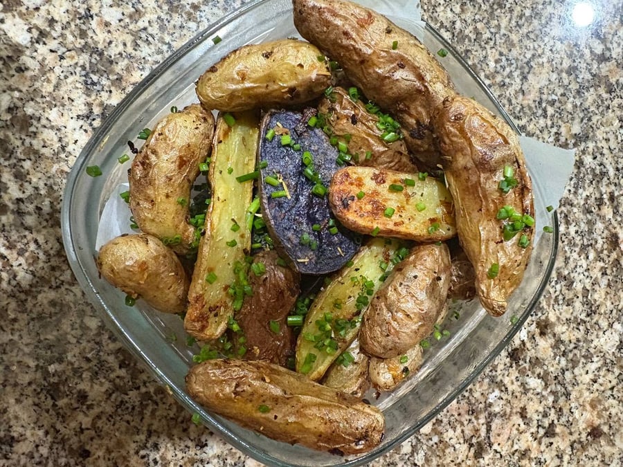 A glass container holds roasted fingerling potatoes with dark purple and gold varieties, garnished generously with freshly chopped bright green chives and fragrant rosemary.