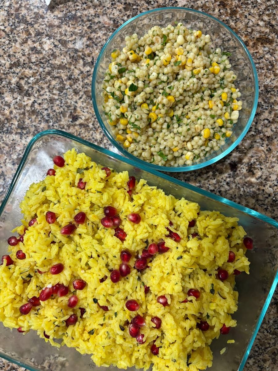 Glass containers holding vibrant saffron basmati rice with pomegranate seeds and pearl couscous mixed with sweet corn and fresh green herbs.