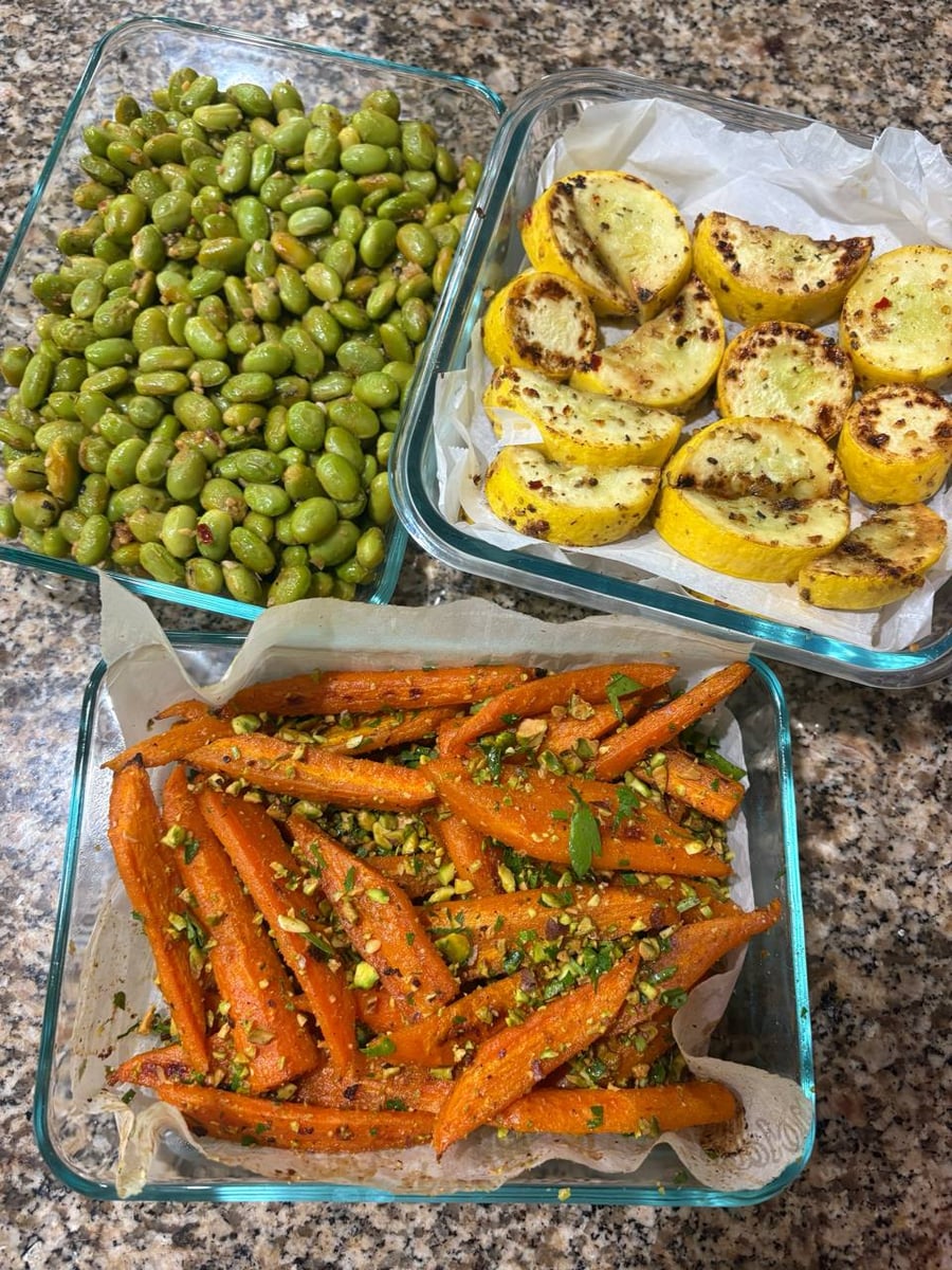 Three glass containers holding charred edamame, pan-seared yellow squash, and coriander roasted carrots topped with crushed pistachios, arranged on a granite countertop.