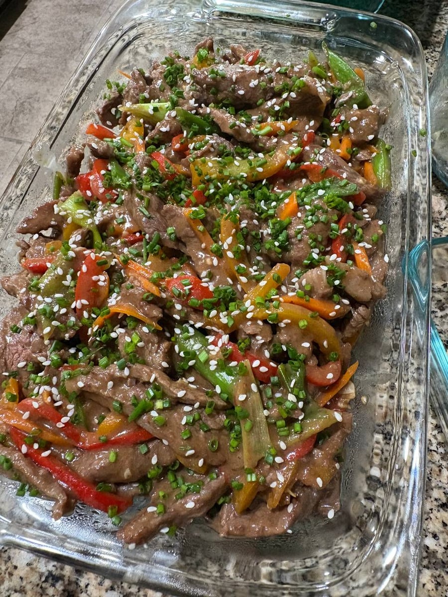 A glass baking dish filled with Mongolian beef, featuring tender steak slices, multicolored bell peppers, sprinkled with sesame seeds and chopped green onions.