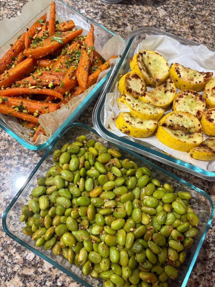 Three glass containers filled with coriander roasted carrots with pistachios, oregano pan-seared yellow squash, and vibrant charred edamame beans with fresh garlic.