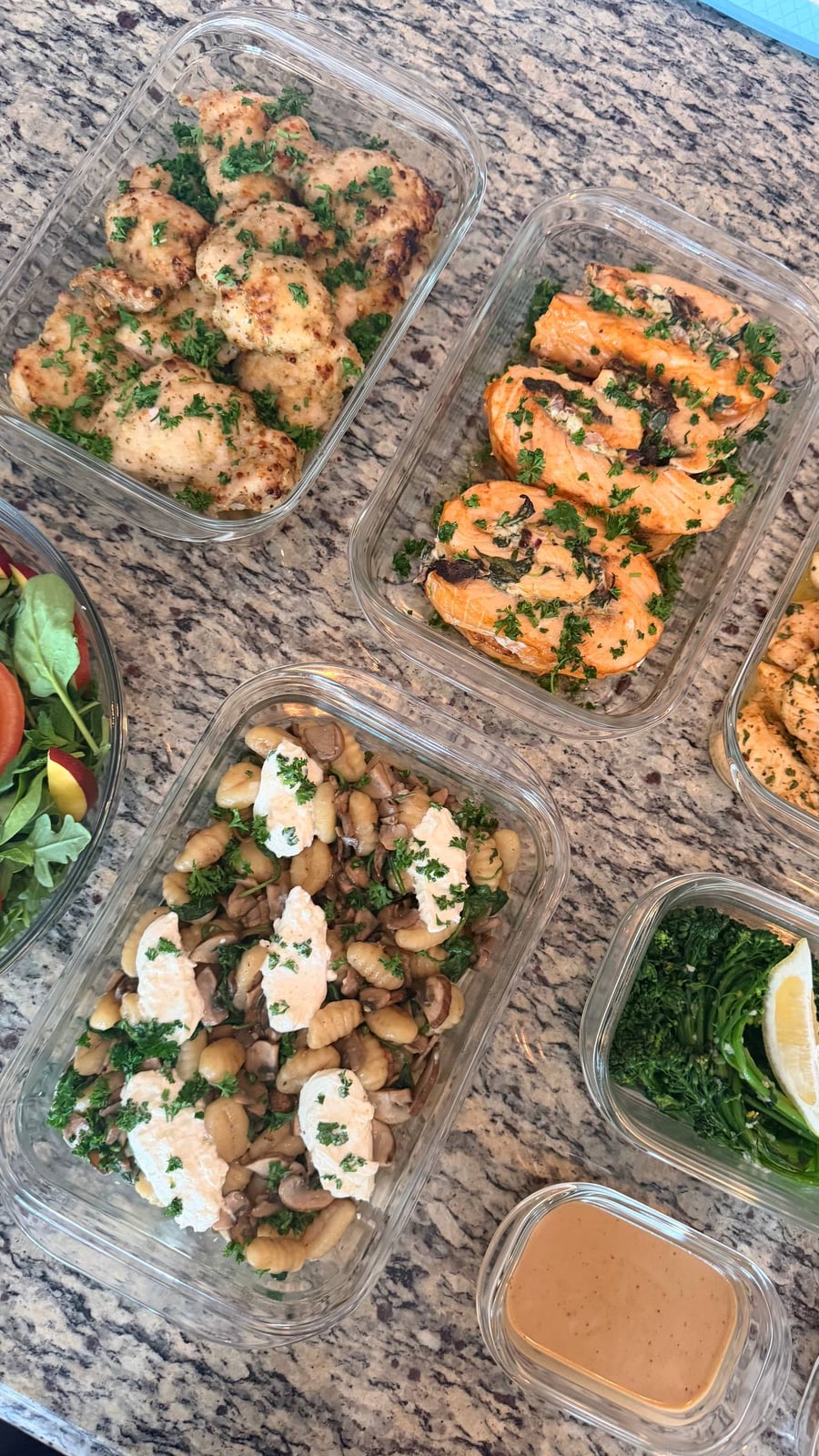 Glass containers featuring garlic parmesan chicken, spinach stuffed salmon pinwheels, mushroom spinach gnocchi with burrata, and lemon garlic broccolini arranged on a stone surface.