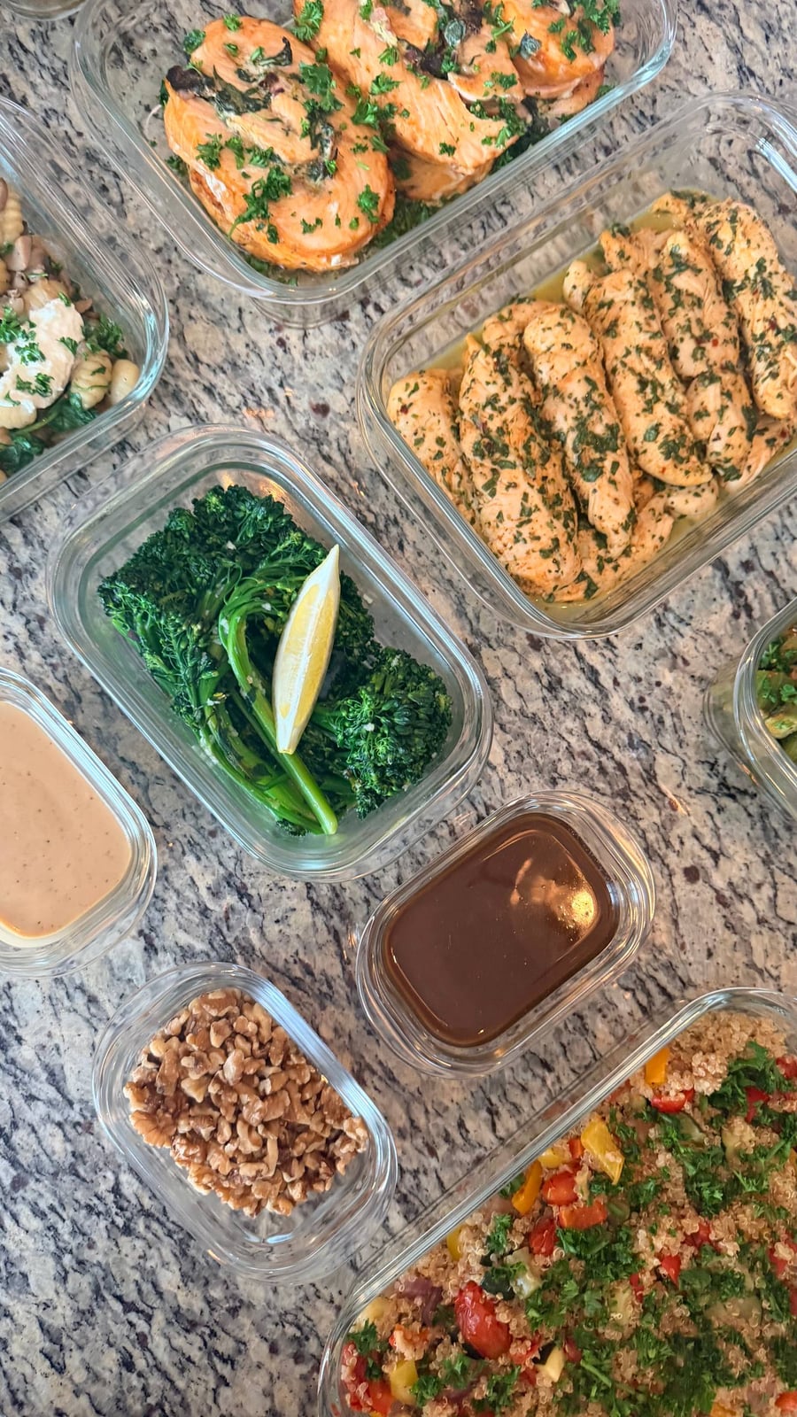 Glass containers displaying spinach stuffed salmon pinwheels, honey lime chicken tenders, lemon garlic broccolini, roasted peppers quinoa, and toasted walnuts on stone surface.
