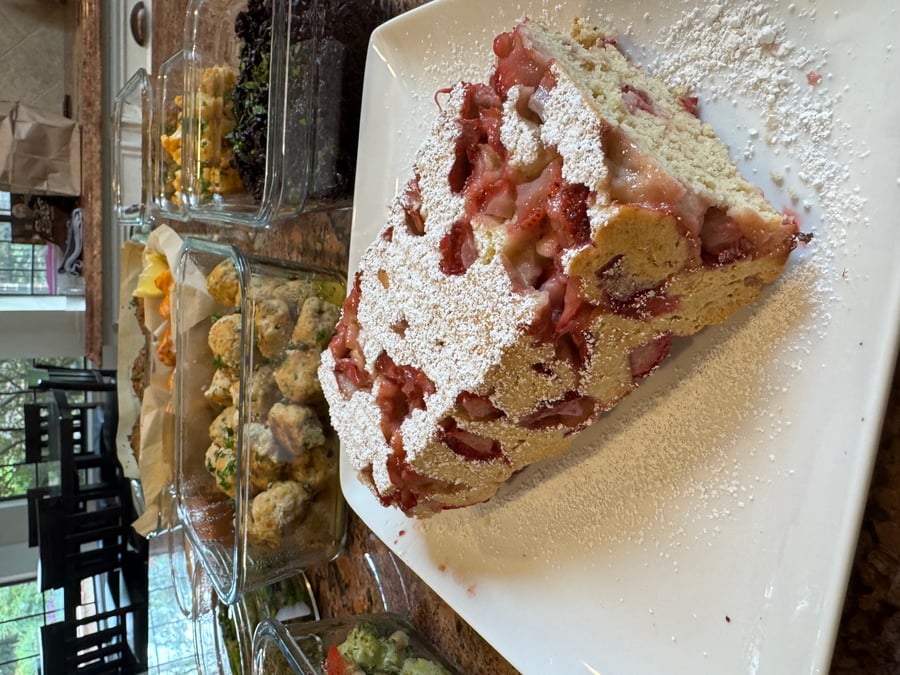 A golden-brown gluten-free strawberry shortcake loaf dusted with powdered sugar, sitting on a square white plate, with meal prep containers in the background.