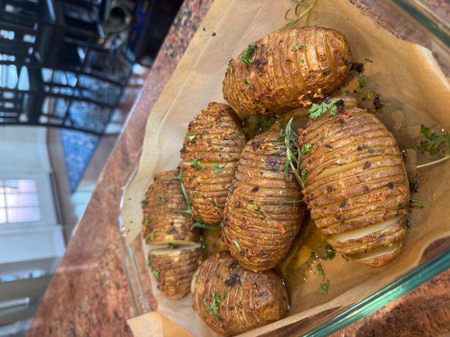 A glass baking dish lined with parchment paper holds several oven-roasted hasselback potatoes seasoned with fresh rosemary, garlic, and bright green parsley.