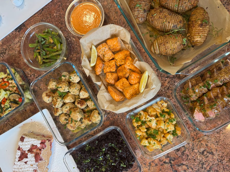 A variety of glass meal prep containers featuring garlic salmon bites, chicken picatta meatballs, pork tenderloin, husselback potatoes, stir-fry vegetables, and a strawberry loaf.