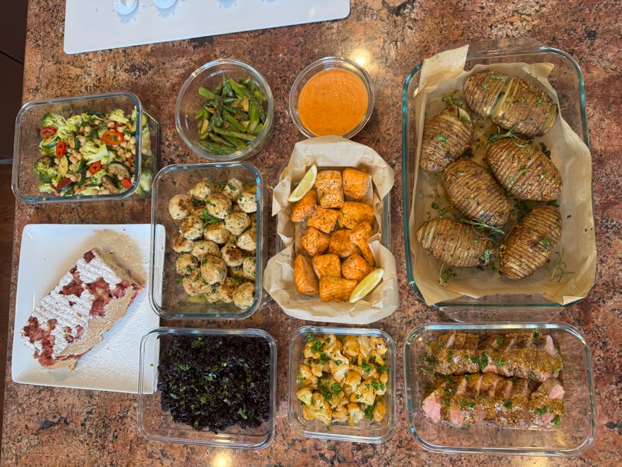 An overhead view of varied meal prep containers featuring garlic salmon, chicken meatballs, pork tenderloin, black rice, roasted potatoes, asparagus, cauliflower, and strawberry loaf.