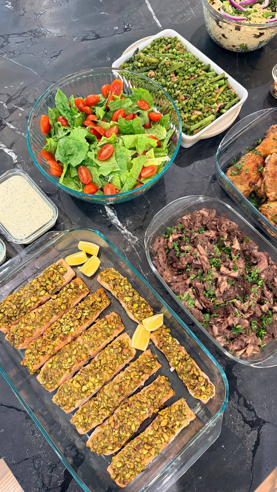 A selection of prepared dishes including pistachio crusted salmon, sauteed beef ribeye with mushrooms, Caesar salad, and seared asparagus served in glass containers.