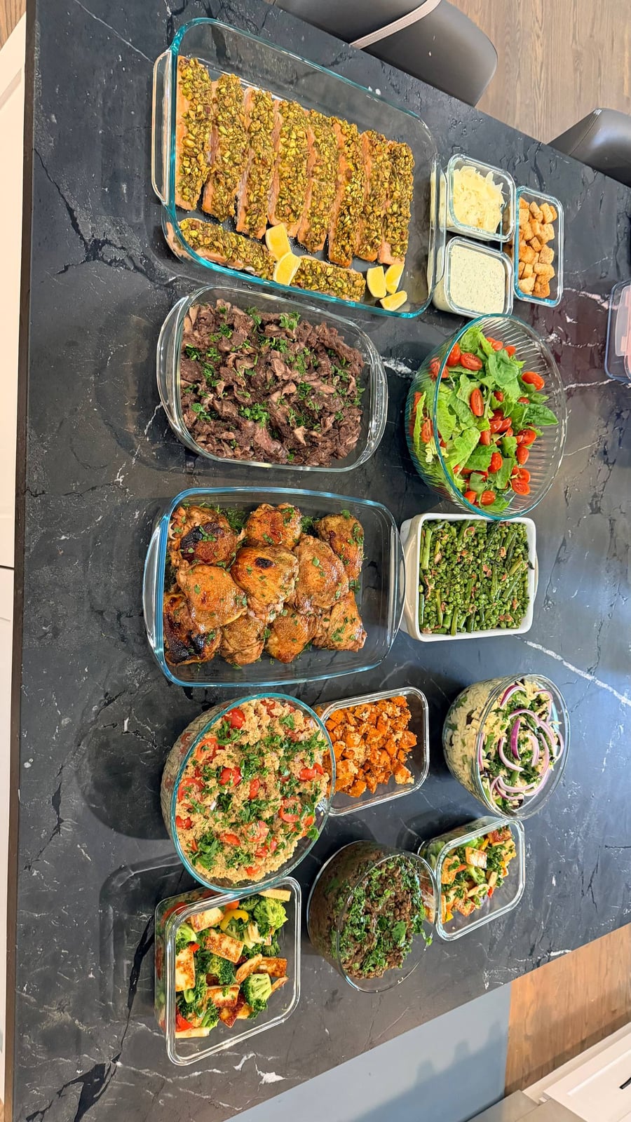 A variety of prepared dishes including pistachio crusted salmon, balsamic chicken, beef ribeye, and quinoa salad arranged on a black marble countertop in glass containers.