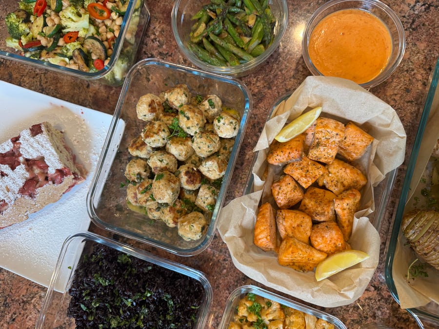 A glass container display of garlic salmon bites, chicken picatta meatballs, cilantro black rice, chili seared broccoli, roasted asparagus, and strawberry shortcake.
