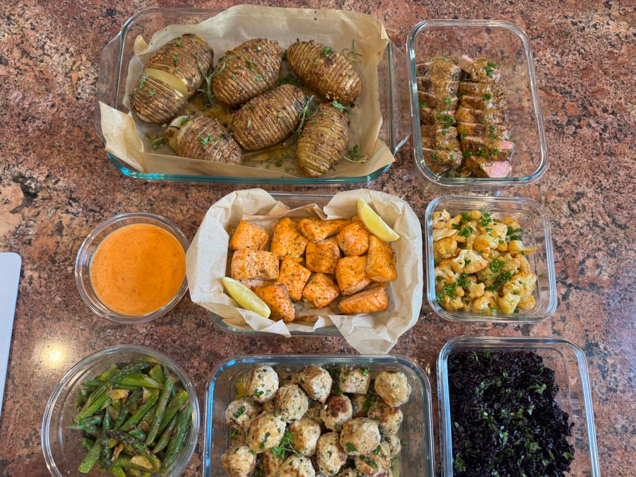 An assortment of glass containers featuring garlic salmon bites, lemon caper chicken meatballs, maple pork tenderloin, rosemary hasselback potatoes, asparagus, cauliflower, and black rice.
