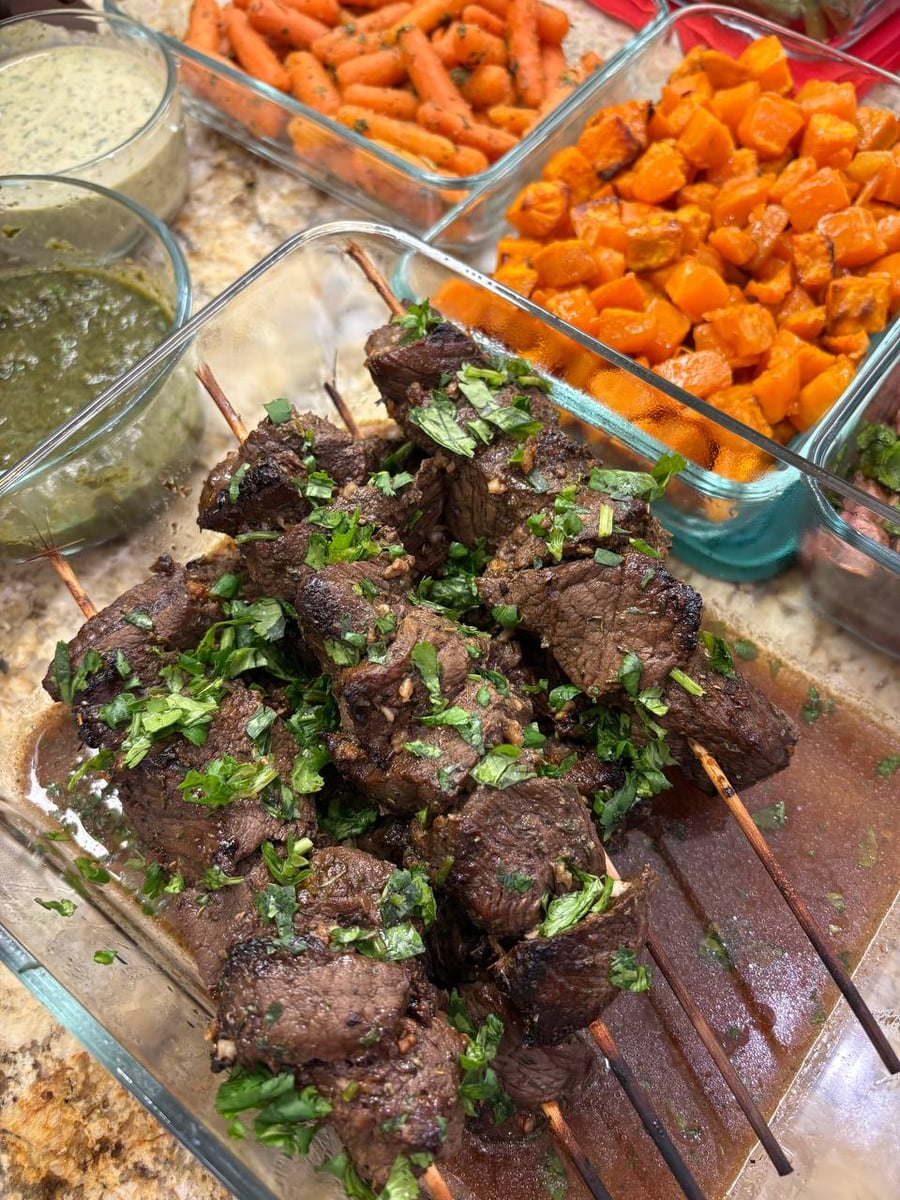 Grilled beef sirloin skewers with fresh herbs served in a glass dish alongside roasted cubed sweet potatoes and two savory herb dipping sauces.