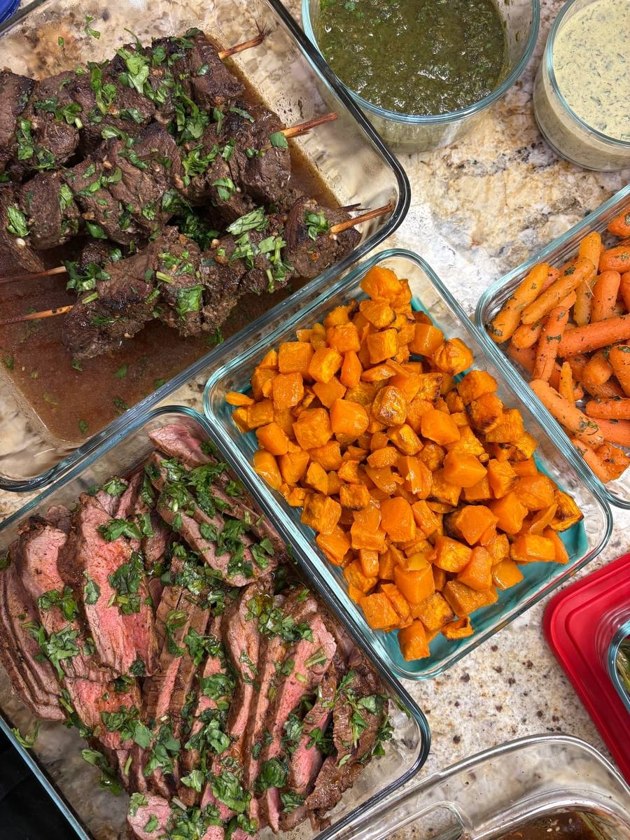Glass containers featuring seasoned beef sirloin skewers, sliced carne asada flank steak, roasted sweet potato bites, and honey glazed carrots with fresh herbs.
