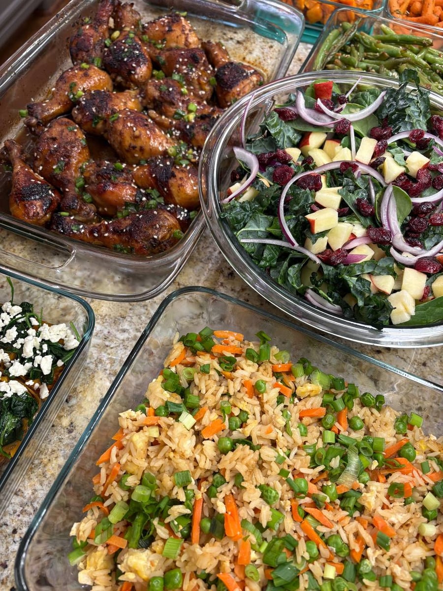 A glass tray of coconut aminos chicken drumsticks, a vibrant kale salad with apples and cranberries, and a glass dish of colorful carrot and pea fried rice.