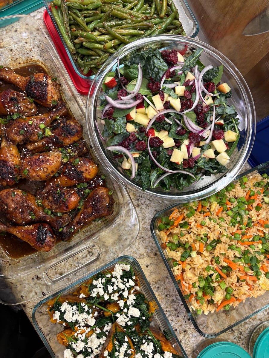 Glass containers featuring coconut amino chicken drumsticks, vibrant carrot and pea fried rice, spinach feta stuffed sweet potatoes, ginger green beans, and kale salad.