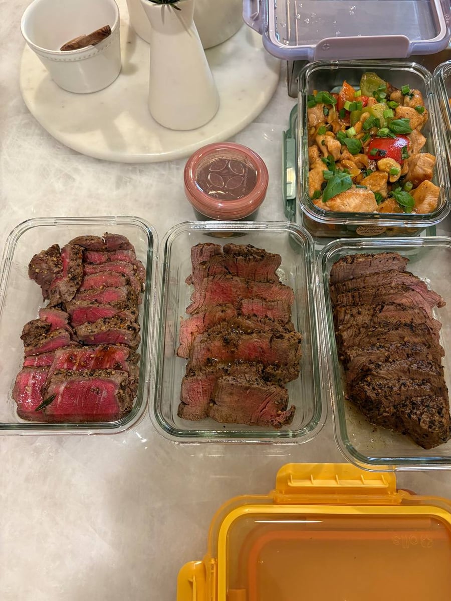 Glass meal prep containers filled with sliced medium-rare rosemary garlic filet mignon and flavorful cashew bell pepper chicken breast bites with fresh green garnishes.