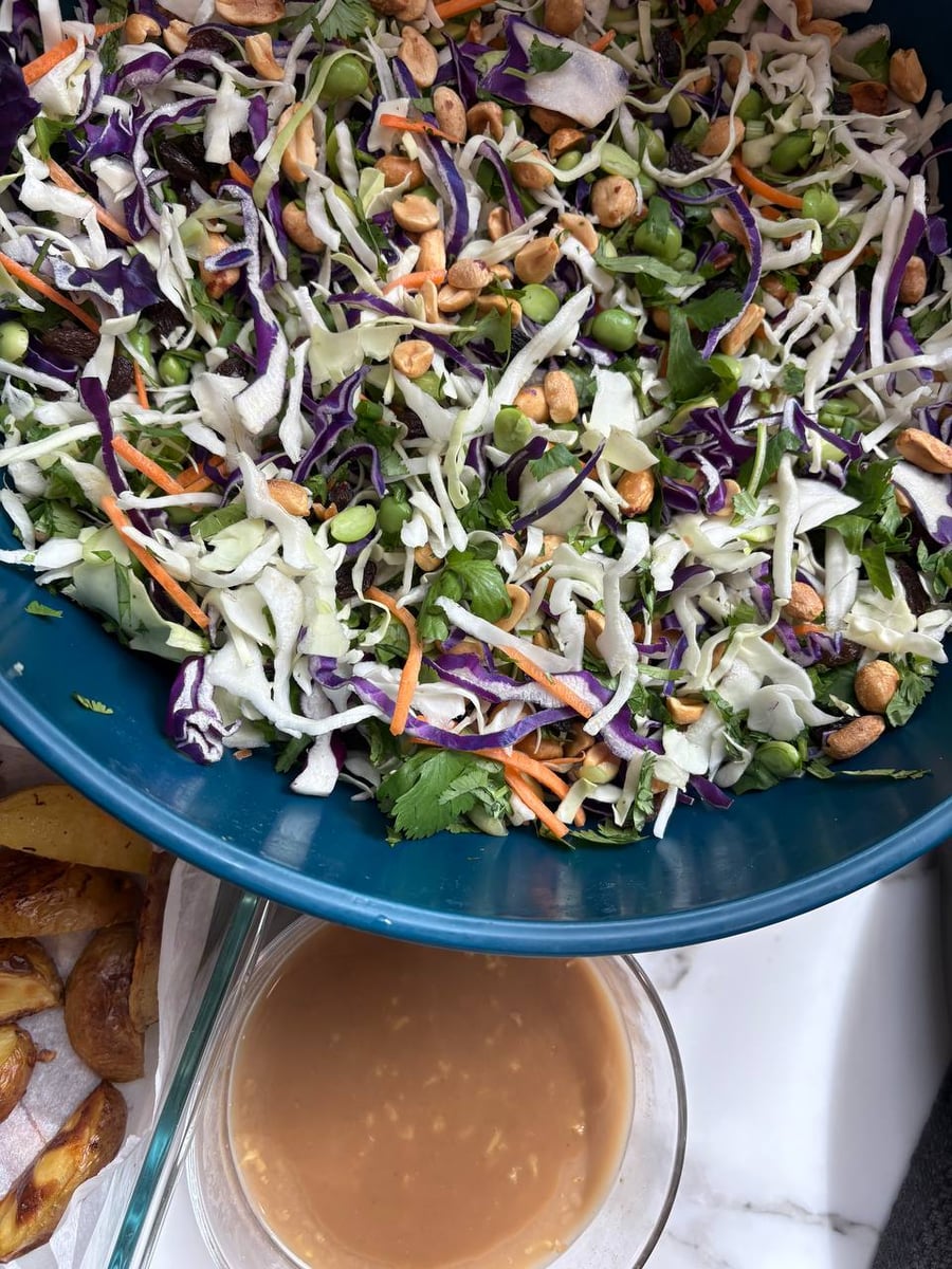 A vibrant Asian-style cabbage salad with edamame, carrots, and peanuts in a blue bowl, served with a side of creamy sesame peanut dressing.