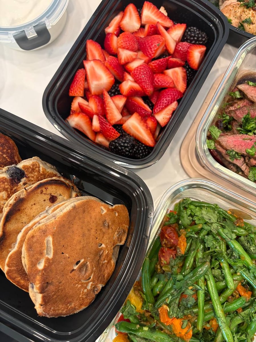 Black and glass containers holding protein blueberry oat pancakes, sliced carne asada flank steak, and vibrant jalapeno green beans with cherry tomatoes.