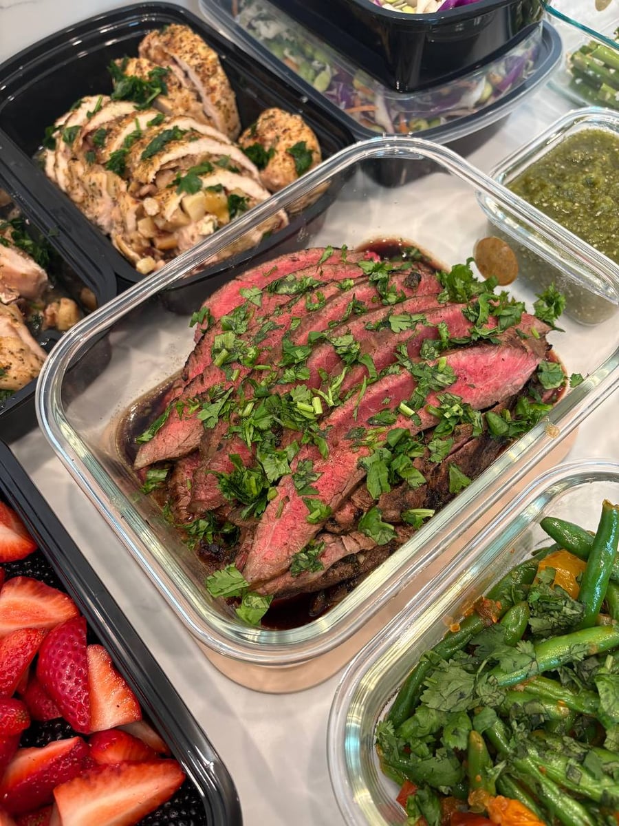 A glass container features sliced medium-rare carne asada flank steak topped with fresh cilantro, positioned alongside containers of turkey roulade and green beans.