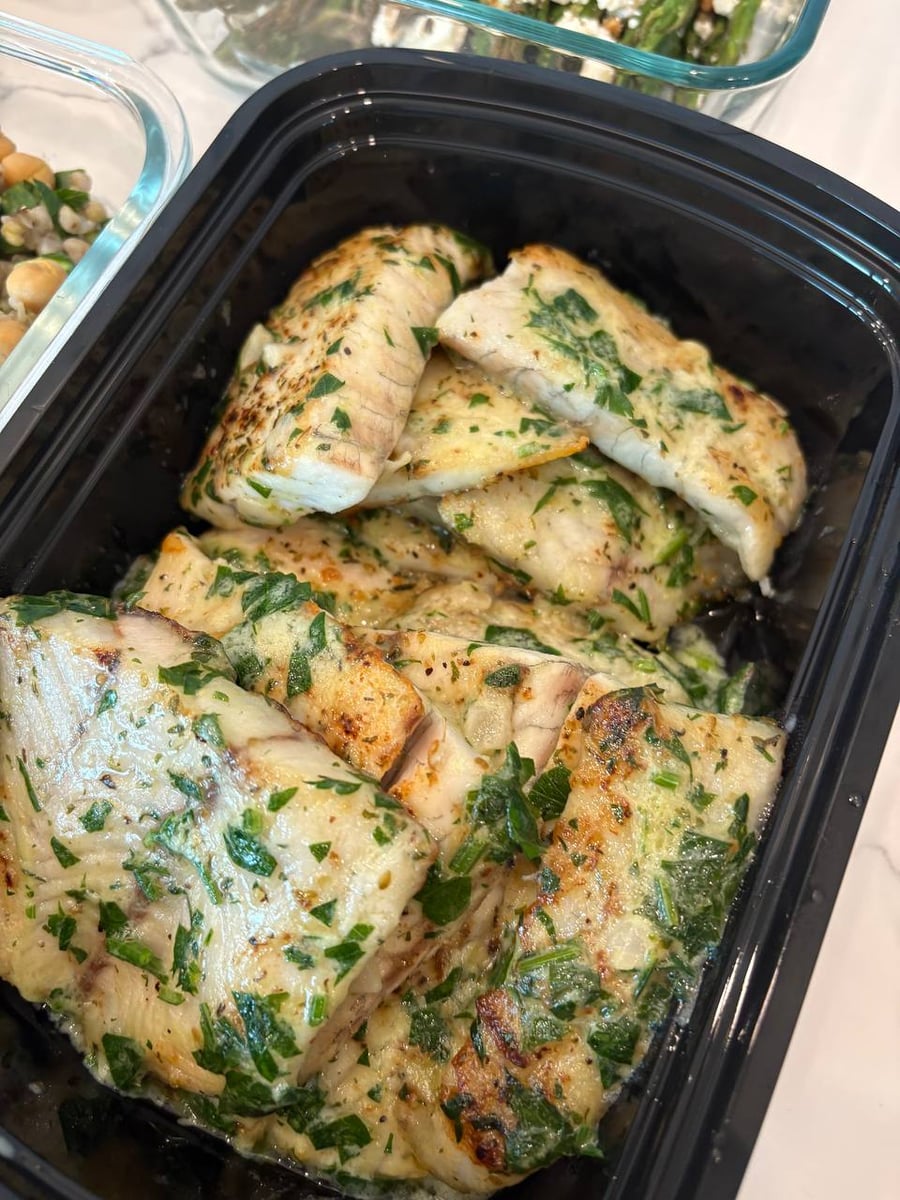 A black plastic container filled with pan-seared barramundi fillets coated in a rich lemon beurre blanc sauce and garnished with fresh green herbs.