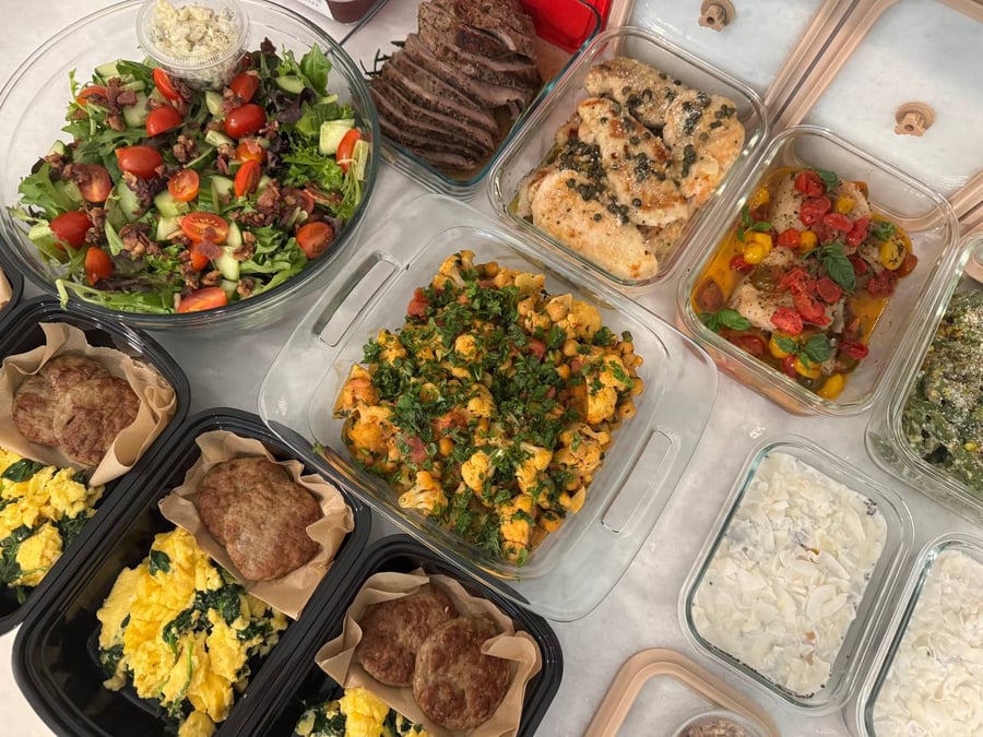 An overhead collection of glass and black meal prep containers featuring roast beef, chicken piccata, tomato basil cod, cauliflower curry, and a loaded chopped salad.