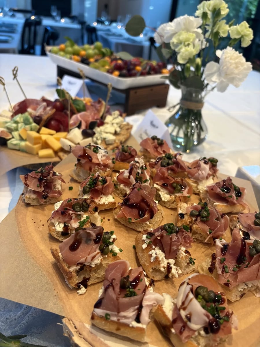 Prosciutto arugula crostini on a wooden board with fruit and cheese boards in the background.
