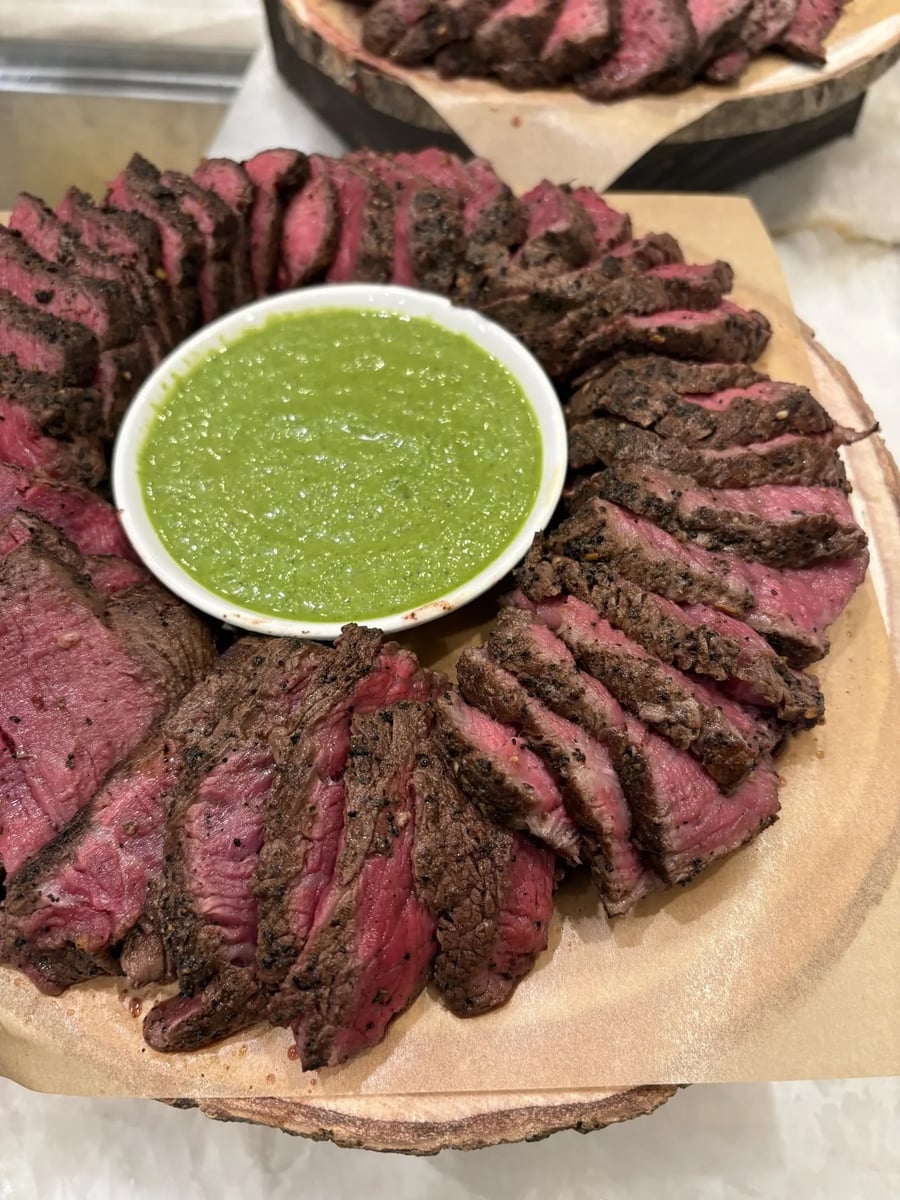 Coffee-crusted beef filet mignon sliced and arranged on a wooden board with Salmoriglio herb sauce.