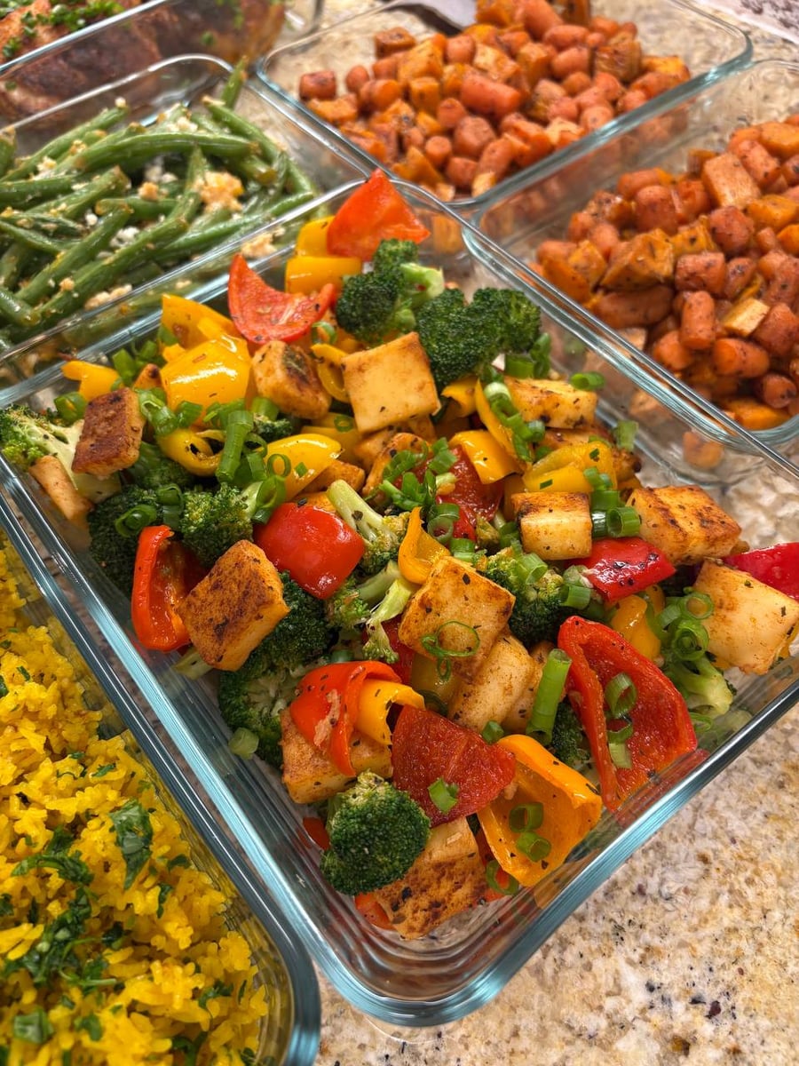 A glass meal prep container filled with colorful bell pepper, broccoli, and paneer stir-fry garnished with chopped green onions on a granite countertop.