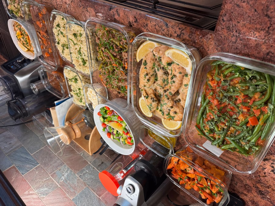 A collection of glass meal prep containers featuring chicken piccata with lemon, sautéed green beans with tomatoes, and roasted sweet potato chunks.