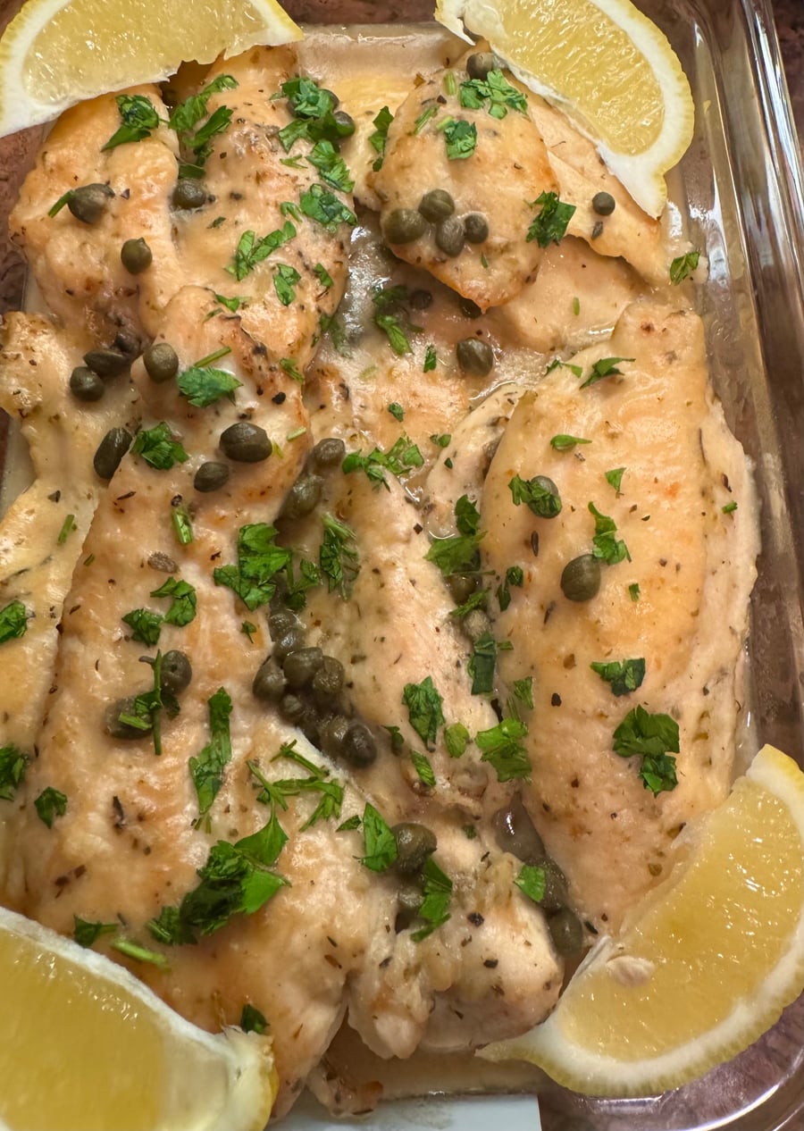 Tender chicken piccata cutlets coated in a silky lemon butter sauce, garnished with briny capers and fresh parsley in a glass baking dish.