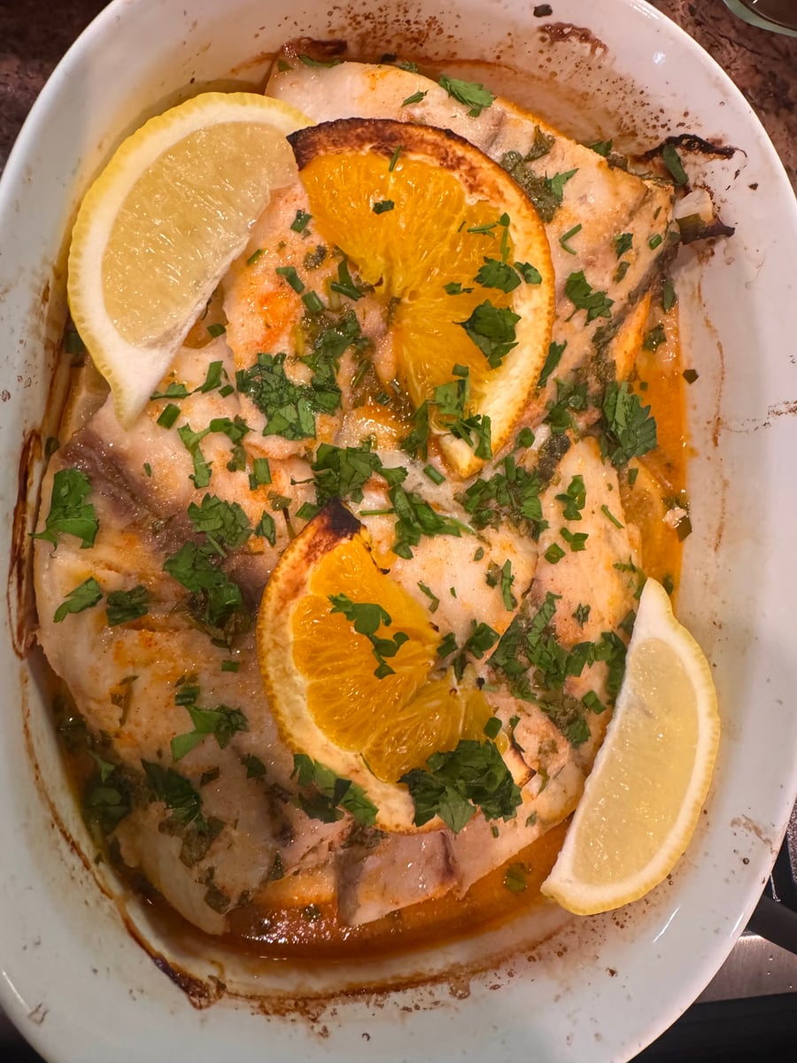 A white oval baking dish filled with tender baked white fish fillets, topped with vibrant orange slices, fresh lemon wedges, and chopped parsley.