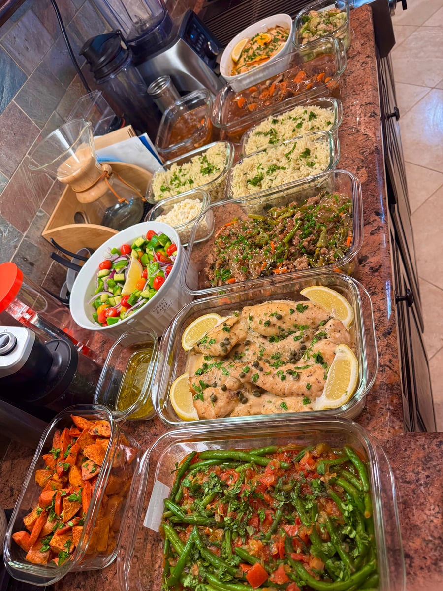 A variety of glass containers filled with chicken piccata, roasted green beans, Mediterranean salad, seasoned rice, and roasted sweet potatoes in a kitchen.