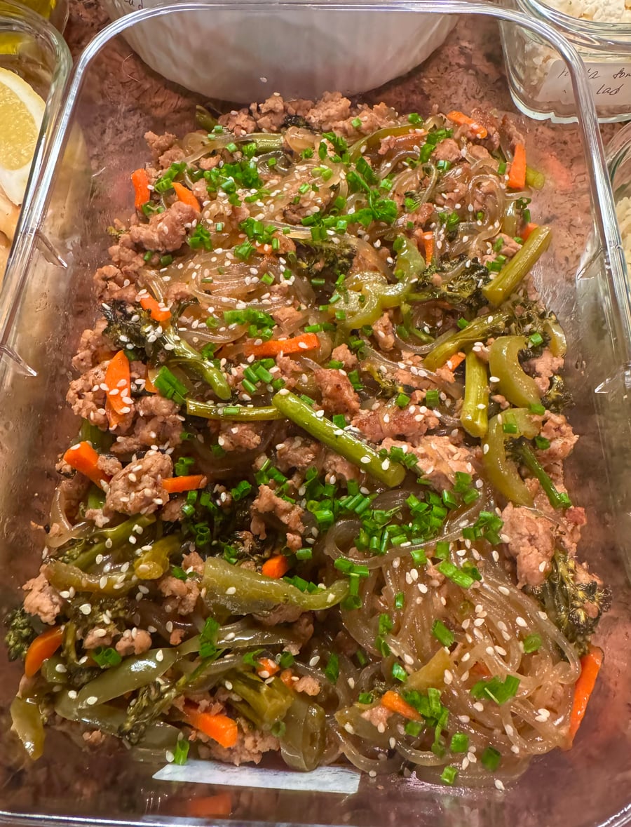 A glass serving dish filled with savory ground pork, translucent glass noodles, stir-fried broccoli, and sliced carrots, all topped with sesame seeds and chives.