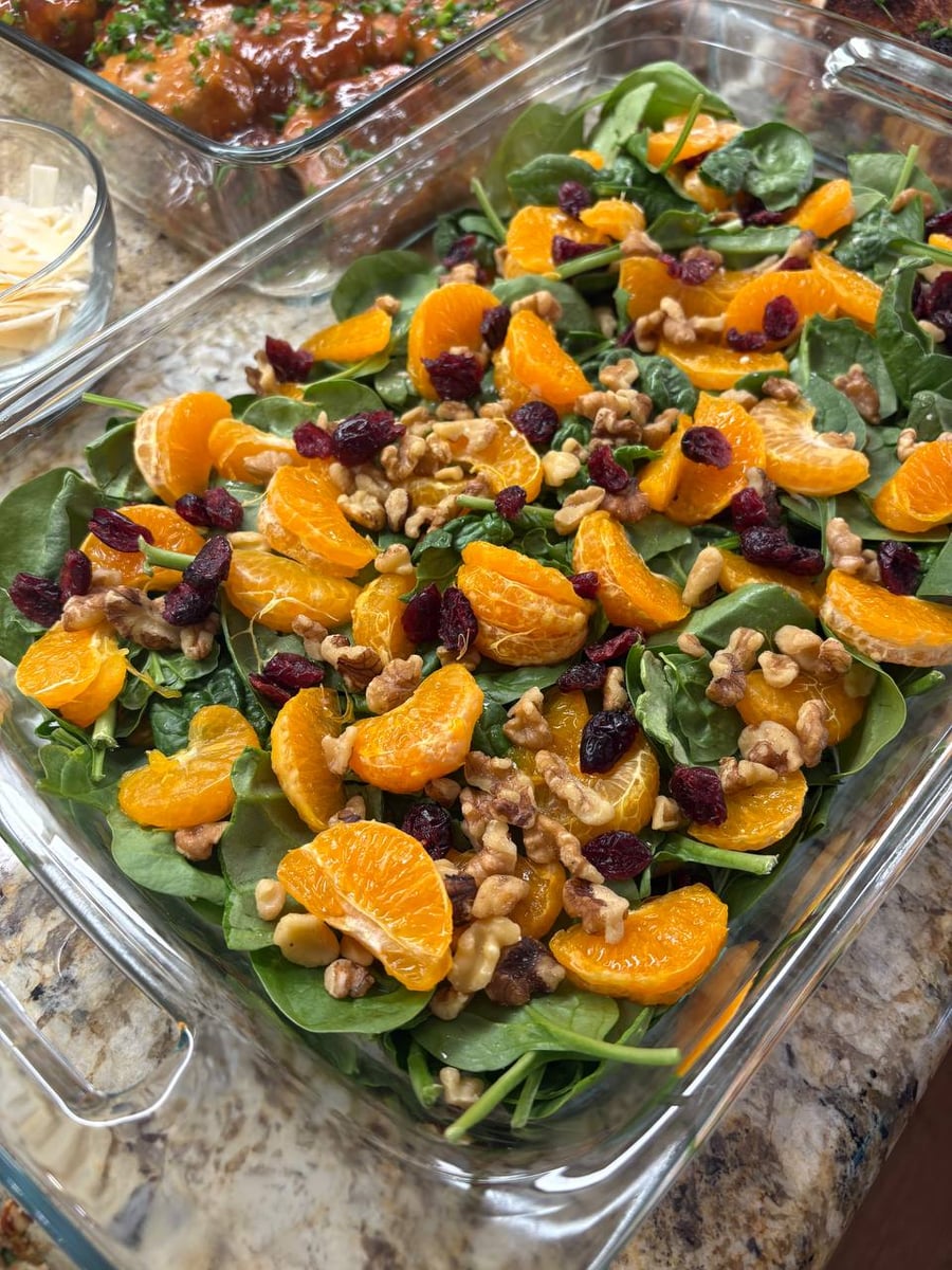 A clear glass rectangular dish holds a vibrant mandarin spinach salad topped with dried cranberries, crunchy walnut pieces, and bright orange segments.