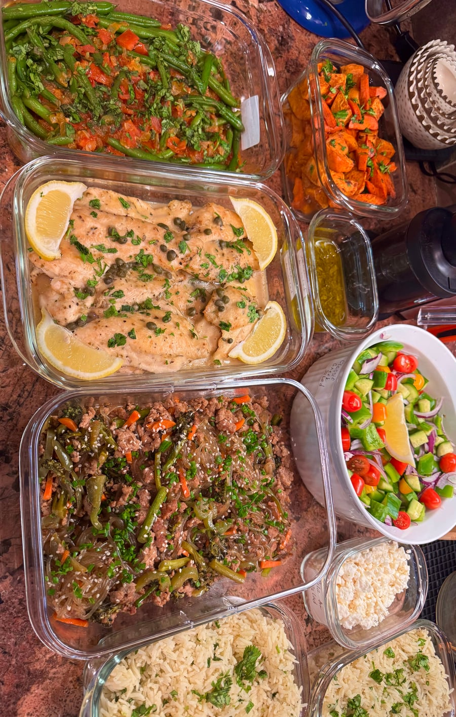 Glass meal prep containers filled with chicken piccata, japchae noodles, green beans, roasted carrots, and a fresh vegetable salad with feta cheese.