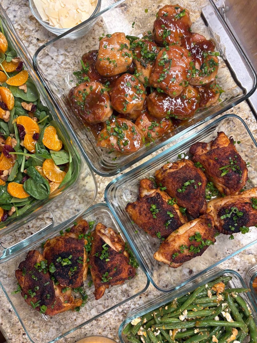 Glass meal prep containers filled with glazed teriyaki pork medallions, spiced paprika chicken thighs, a fresh mandarin spinach salad, and garlic goat cheese beans.