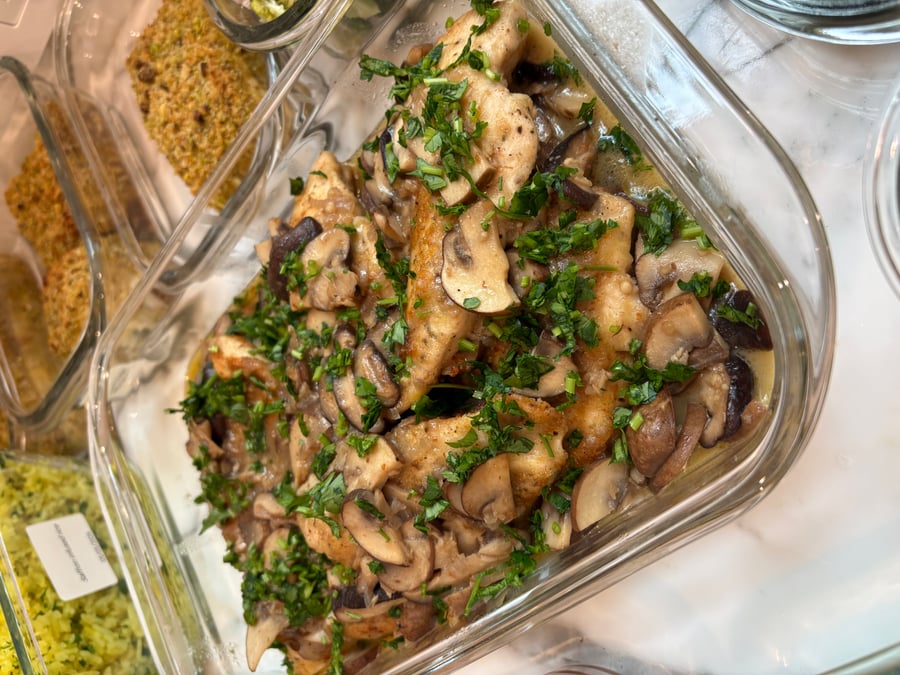 Glass meal prep container featuring tender chicken breast Marsala topped with sautéed mixed mushrooms and freshly chopped bright green parsley garnish.
